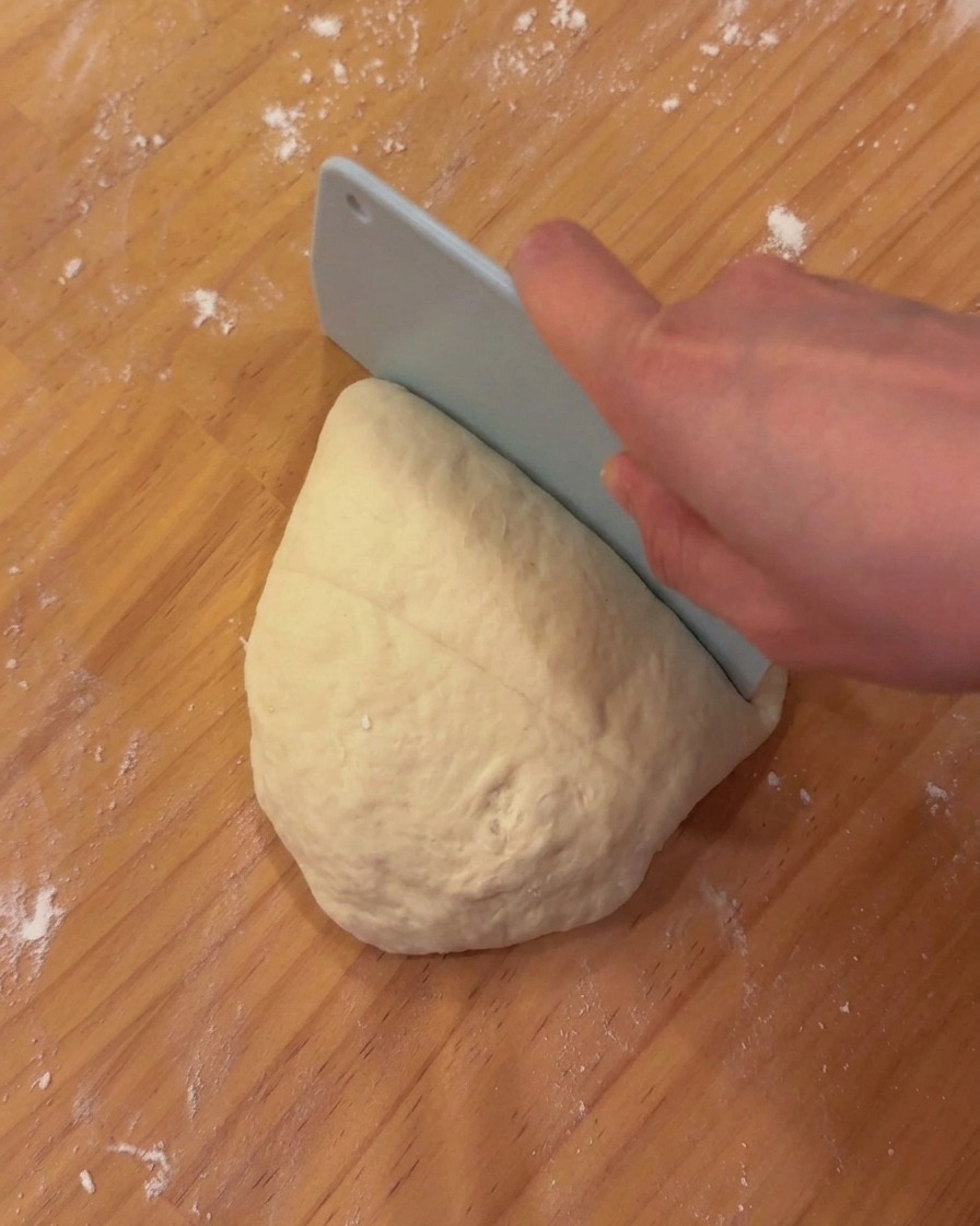 Porciones de masa de pizza suave y fermentada reposando sobre una tabla de madera junto a una rasqueta de panadero.
