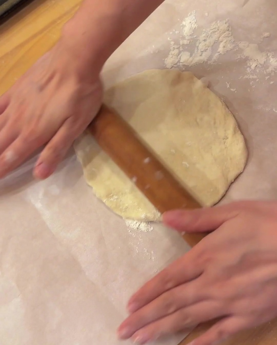 Un rodillo de madera aplanando una pieza redonda de masa de pizza sobre papel de horno.