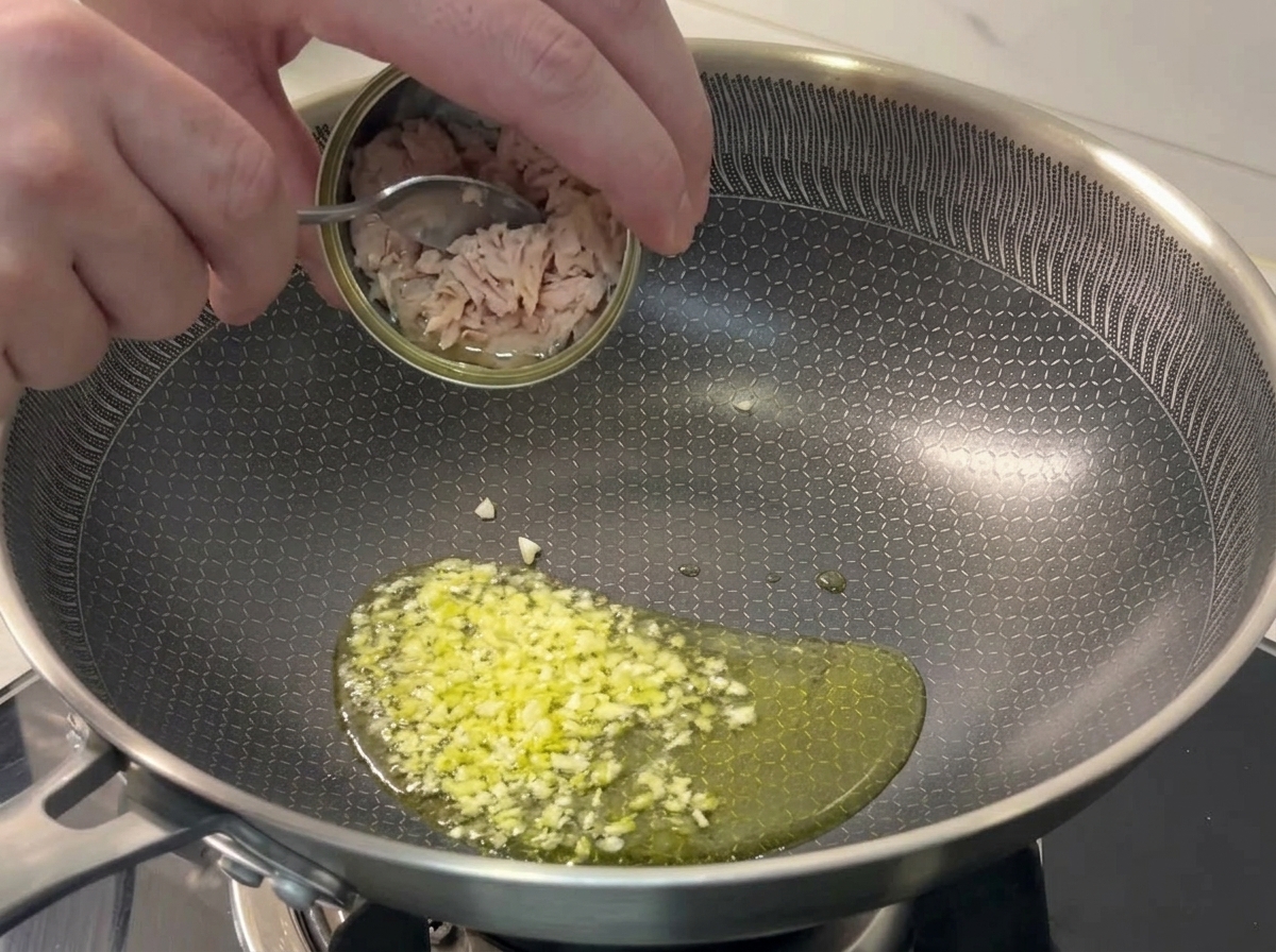 Cuchara añadiendo trozos de atún en una sartén con ajo sofrito.