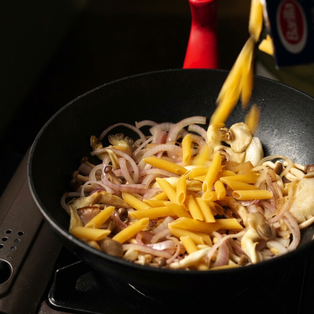 Pasta penne seca vertiéndose de una caja en una sartén negra con champiñones y cebollas salteados.