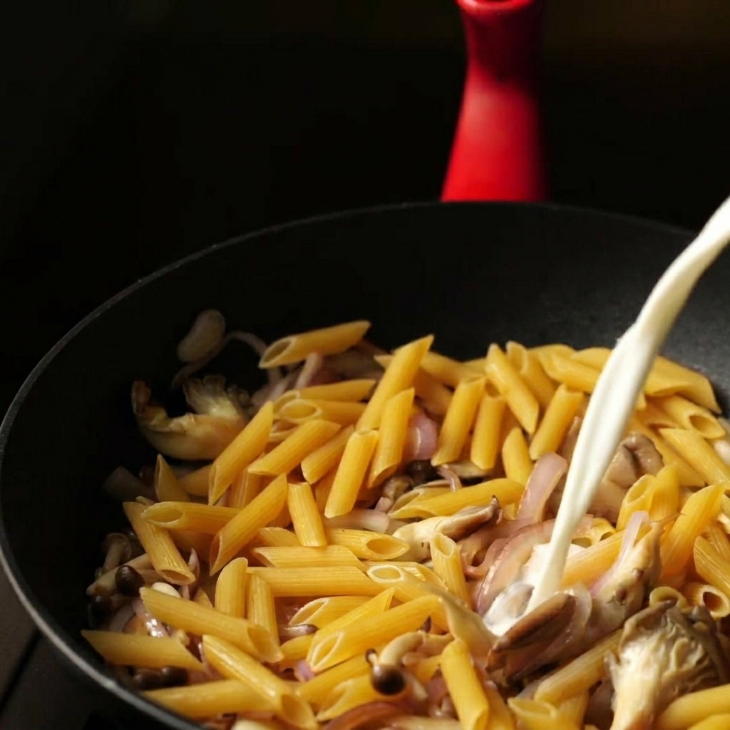 Un chorro constante de mezcla de leche y agua vertido en una sartén sobre penne seco y verduras salteadas.