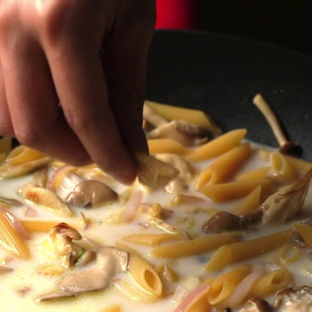 Dedos dejando caer un cubo de caldo en una sartén llena de pasta penne cocinándose en un caldo blanco cremoso.