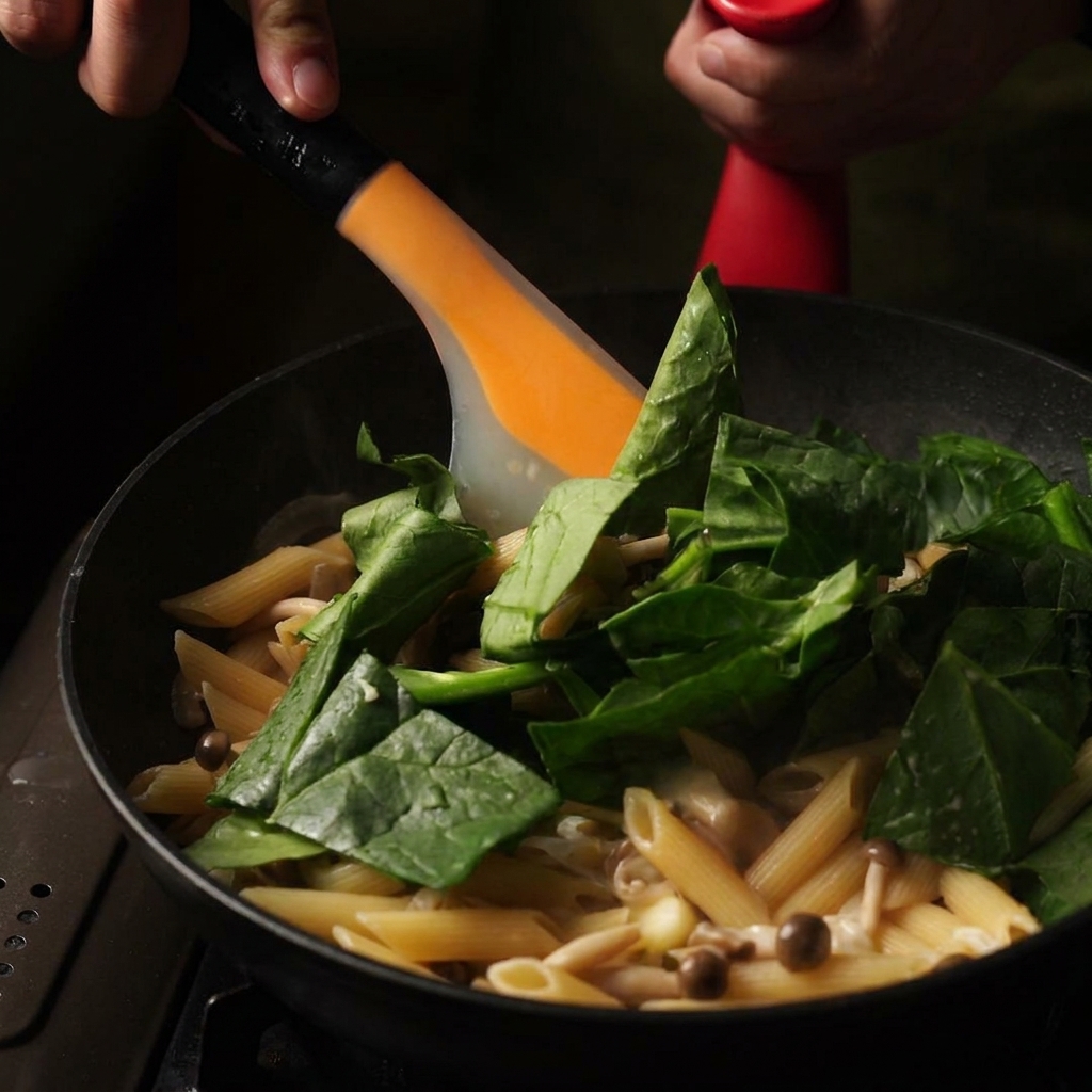 Una espátula naranja revolviendo hojas de espinaca verde fresca y mantequilla en una sartén de penne cremoso cocido.