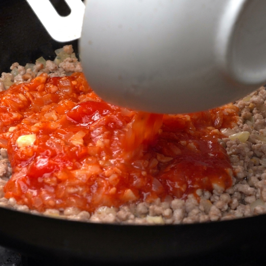 Tomates triturados vibrantes siendo vertidos desde un cuenco blanco en una sartén con carne picada cocinada.