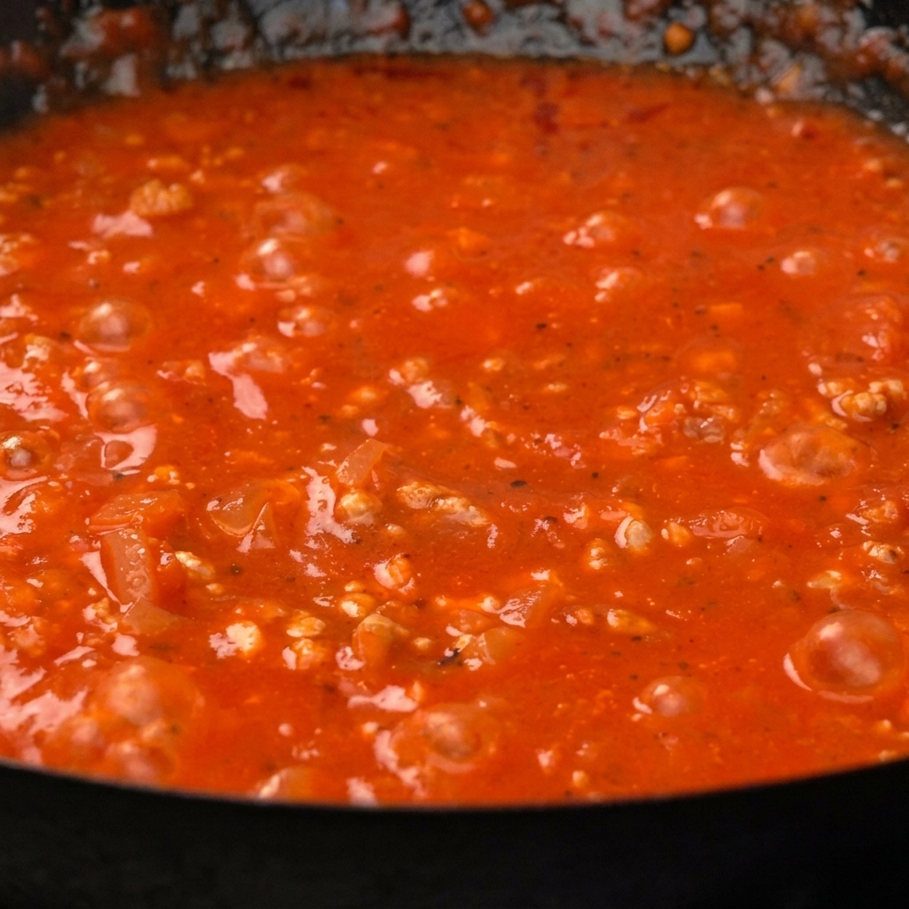 Pasta paccheri cocida siendo mezclada en una espesa y vibrante salsa de carne y tomate en una sartén.