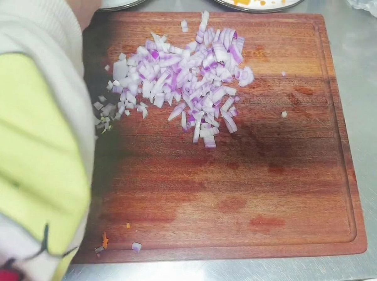 El brazo de una persona cortando una cebolla roja en dados pequeños sobre una tabla de cortar de madera.