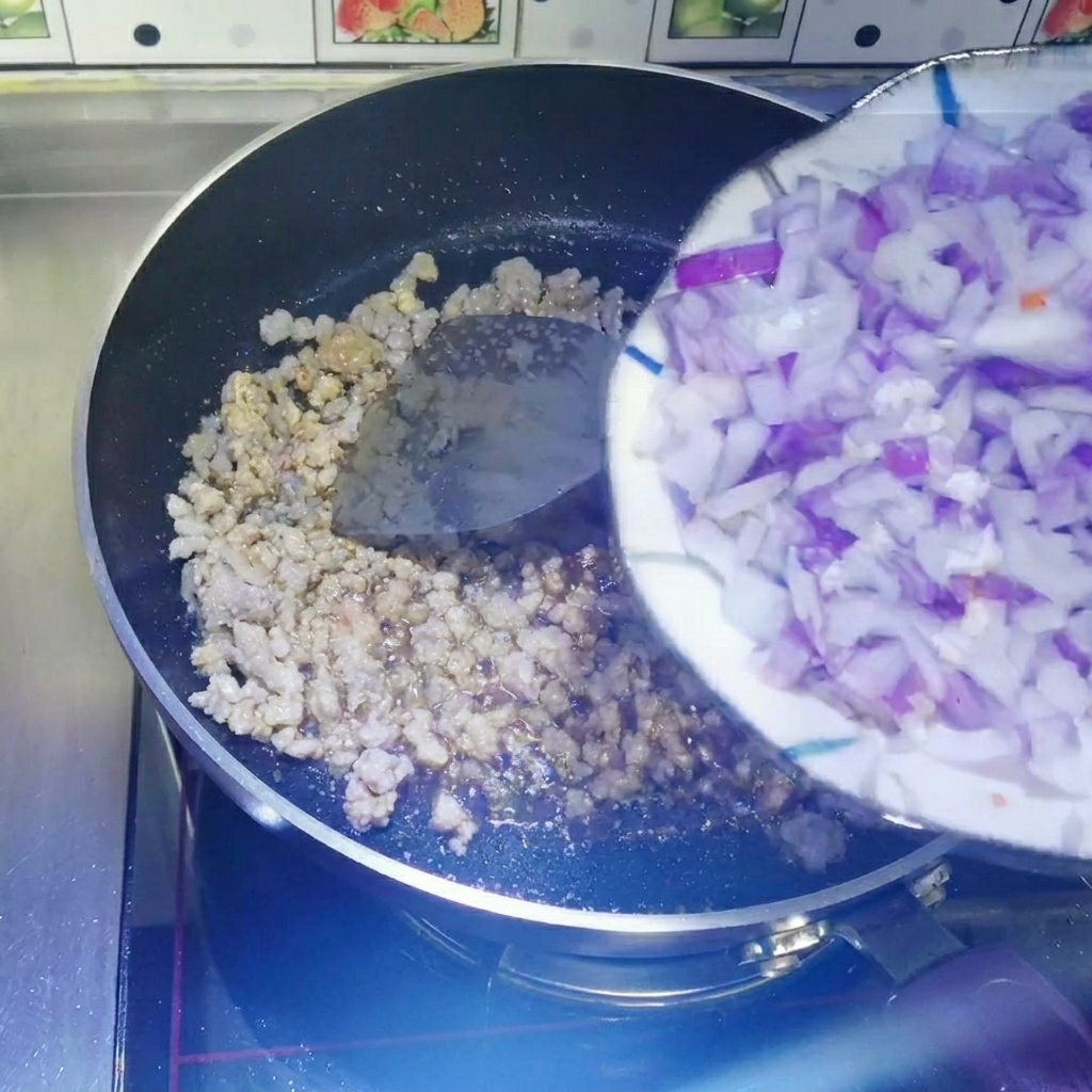 Cebollas rojas picadas siendo arrastradas desde un plato blanco hacia una sartén antiadherente con carne picada cocida.