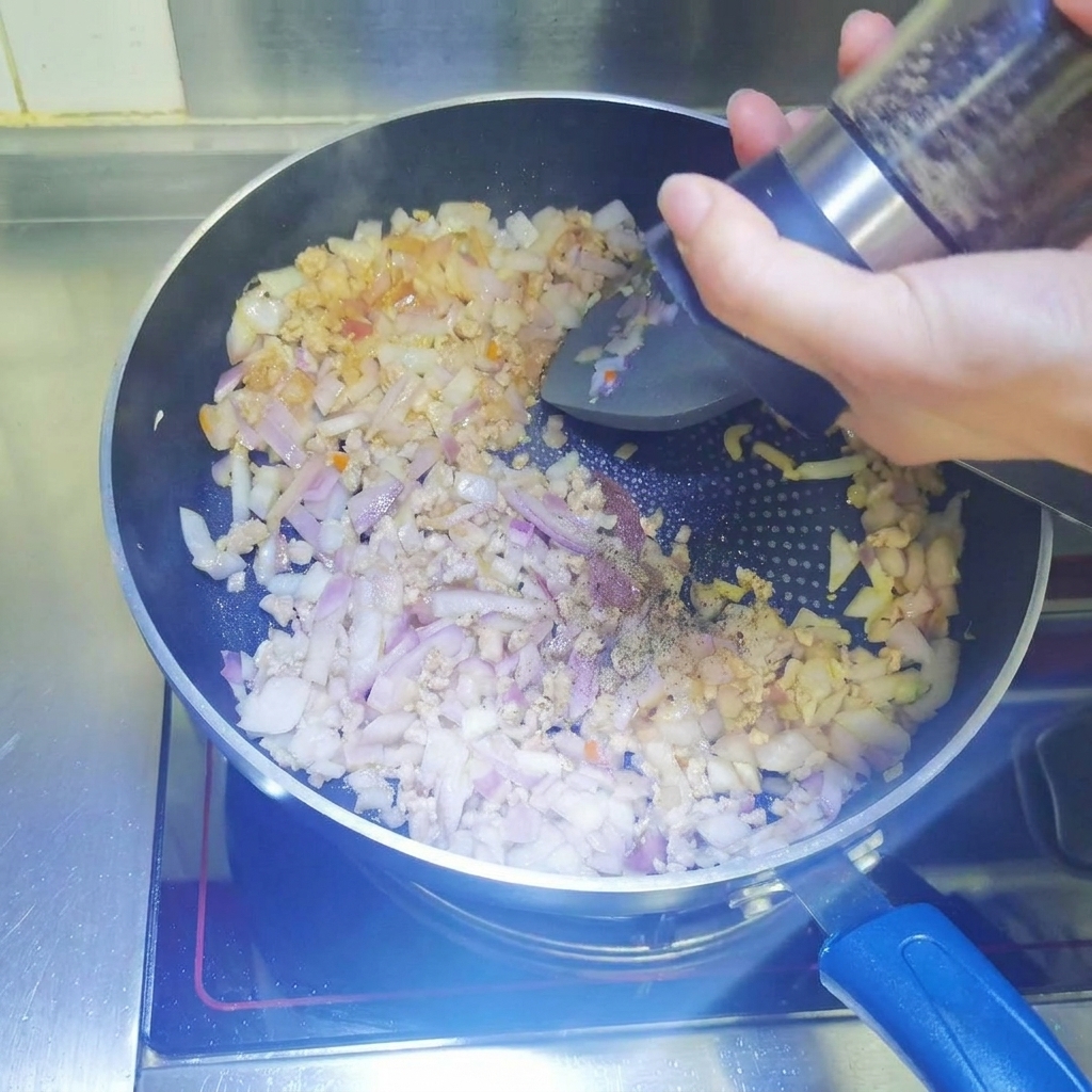 Pimienta negra siendo molida fresca desde un molinillo hacia una sartén con carne picada y cebollas.