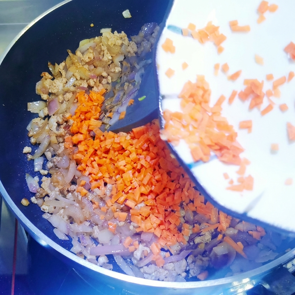 Zanahorias picadas siendo añadidas a una sartén con carne y cebollas salteadas, mezcladas con una espátula de silicona.