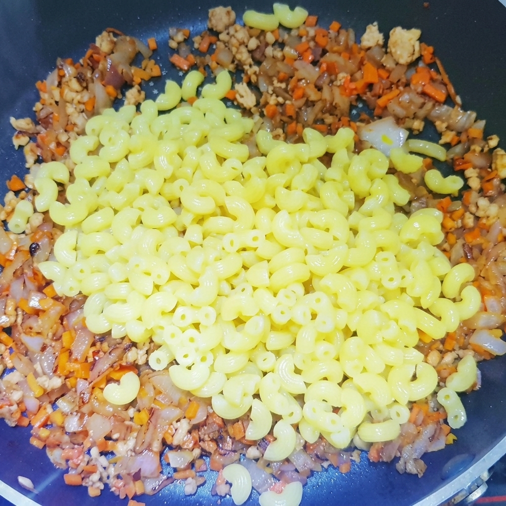 Macarrones hervidos siendo añadidos a una sartén llena con una base de carne picada, cebollas y zanahorias.
