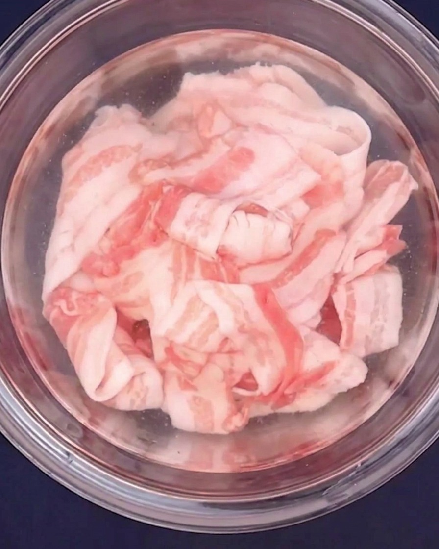 Panceta de cerdo cortada en finas rodajas siendo escaldada con agua caliente en un bol de cristal.