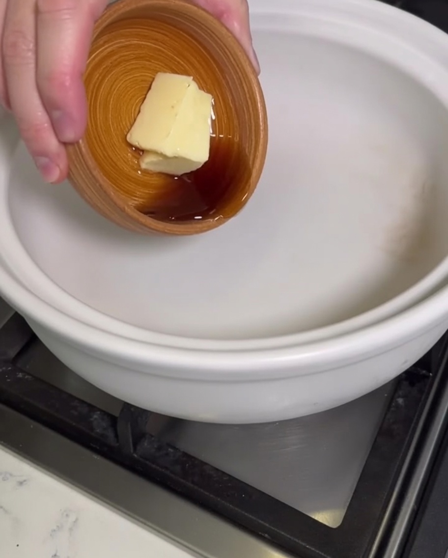 Una mano añadiendo un bloque de mantequilla y aceite de sésamo líquido a una olla de barro blanca.