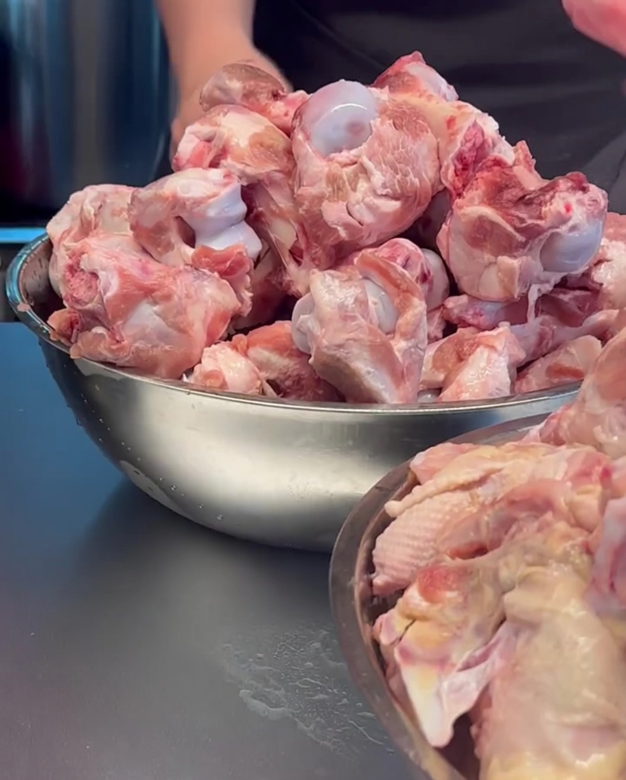 Huesos de cerdo crudos y carcasas de pollo siendo colocados en una olla metálica grande.