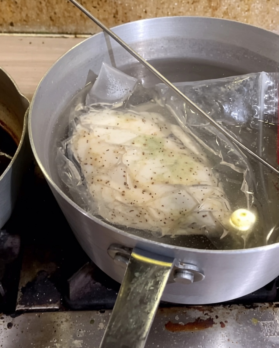 Una bolsa sellada al vacío que contiene pechuga de pollo sazonada sumergida en una olla de agua caliente.