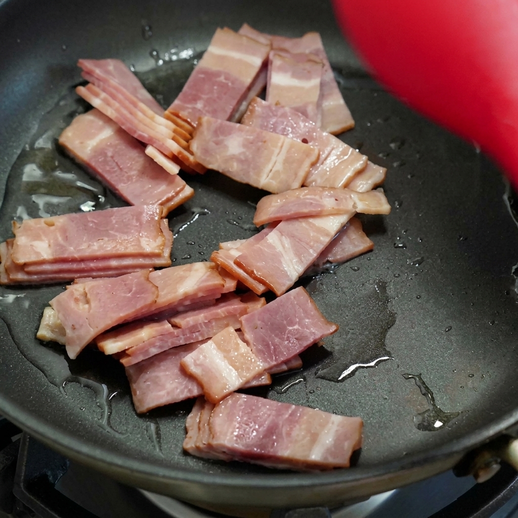 Tiras de bacon cocinándose y dorándose en una sartén negra, siendo removidas con una espátula roja.