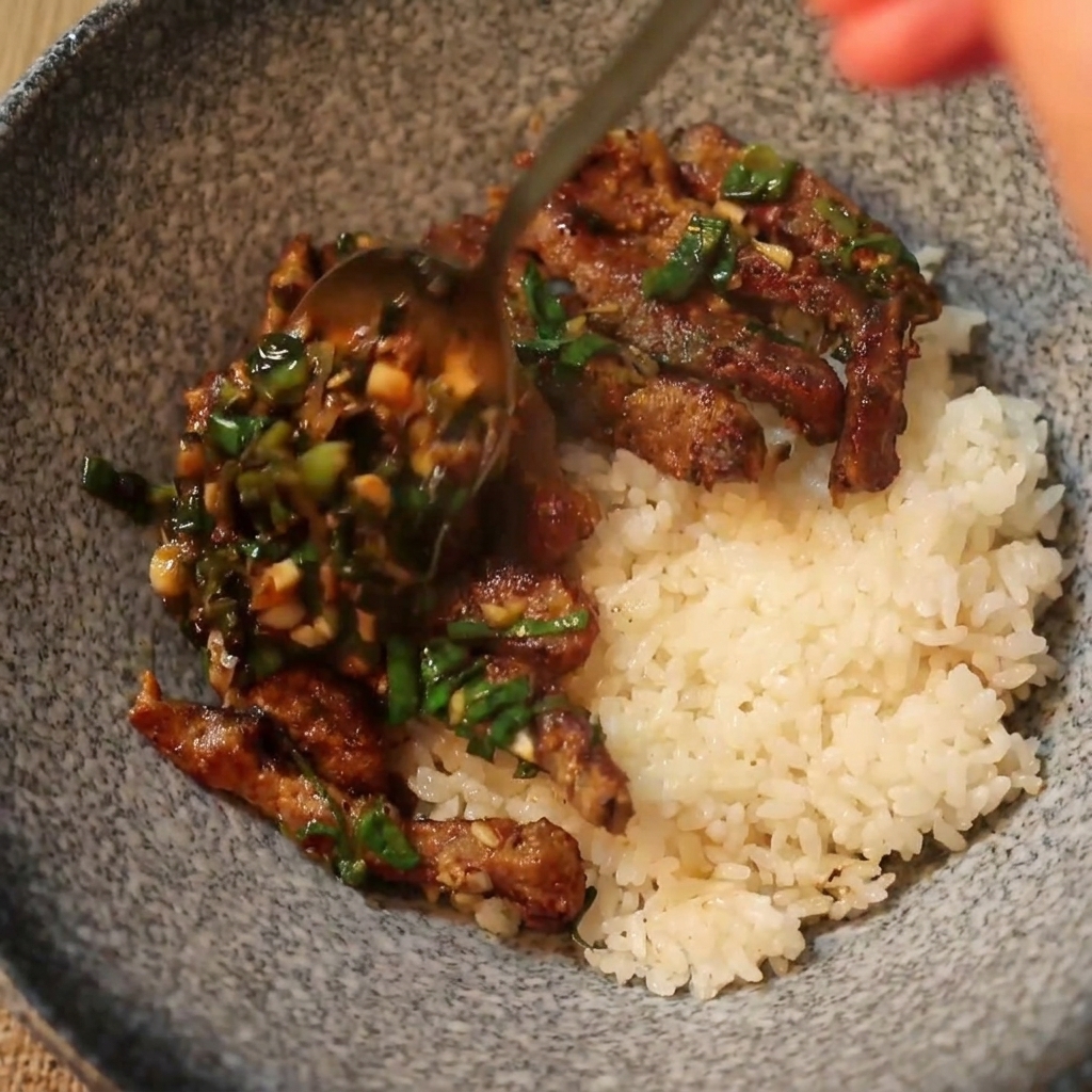 Una cuchara sirviendo ternera caliente cocinada y salsa de cebollino sobre una cama de arroz blanco en un cuenco de cerámica.