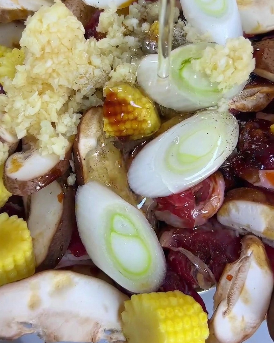 Aceite de cocina vertiéndose sobre una bandeja que contiene rodajas de ternera, champiñones shiitake, maíz baby, cebollines y ajo.
