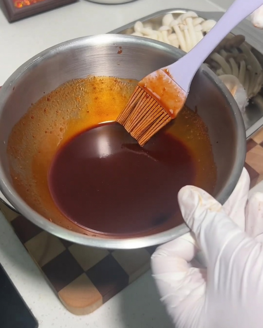 Un tazón de metal que contiene una mezcla de salsa oscura y picante siendo revuelta con un pincel de silicona.