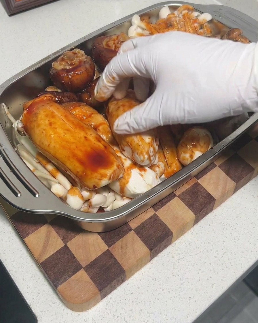 Manos con guantes masajeando la salsa sobre las setas en una bandeja para asegurar una cobertura completa.