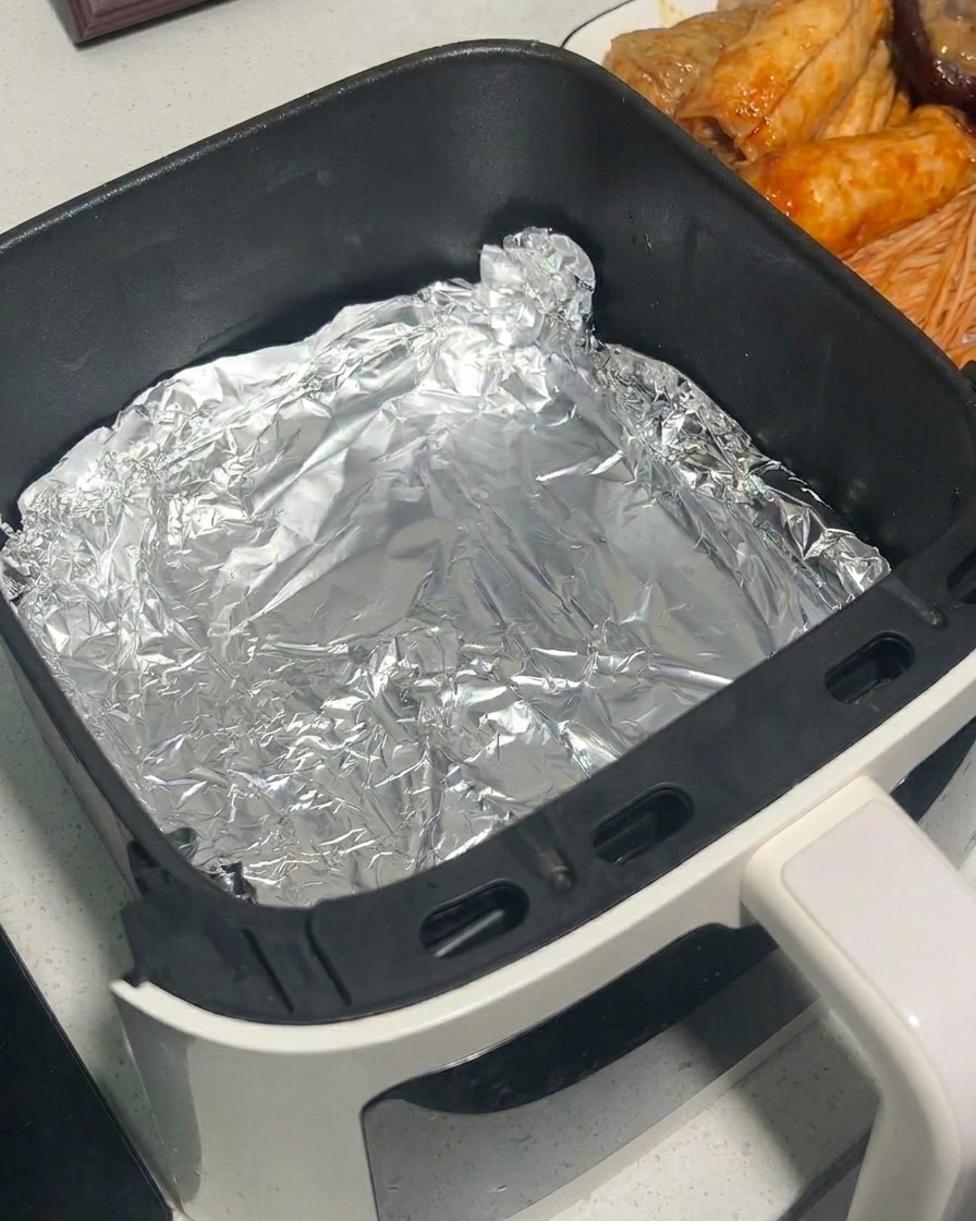 Setas marinadas siendo colocadas en la canasta de una freidora de aire forrada con papel de aluminio usando pinzas de metal.