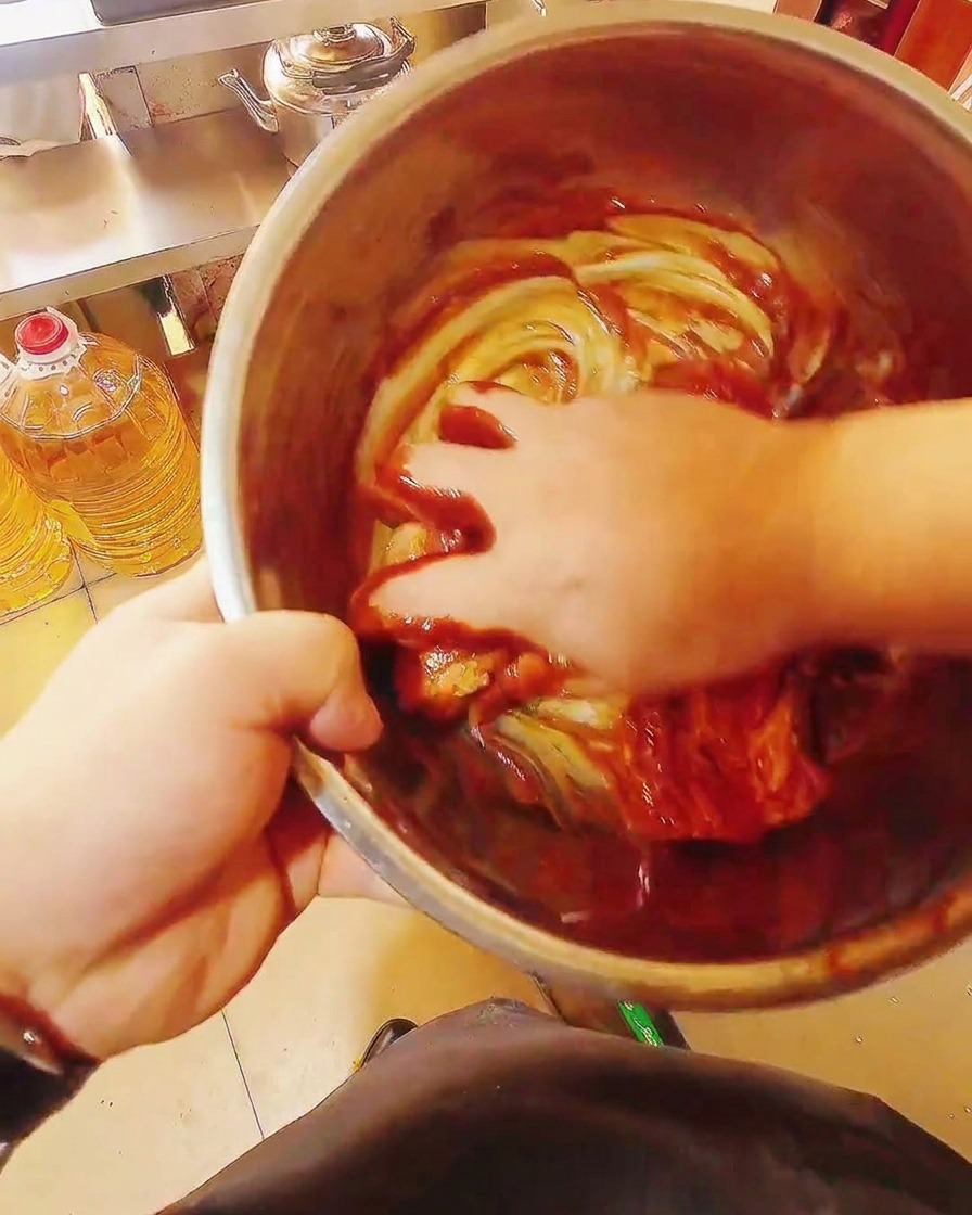 Tiras de panceta de cerdo crudas mezclándose a mano en un bol con un marinado rojo espeso y brillante.