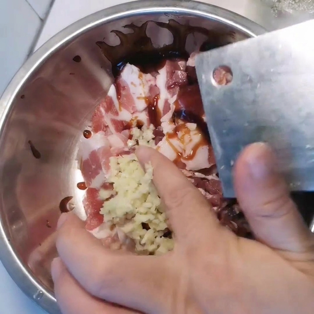 Un bol de metal que contiene la panceta de cerdo en rodajas siendo sazonada con ajo picado y varias salsas.