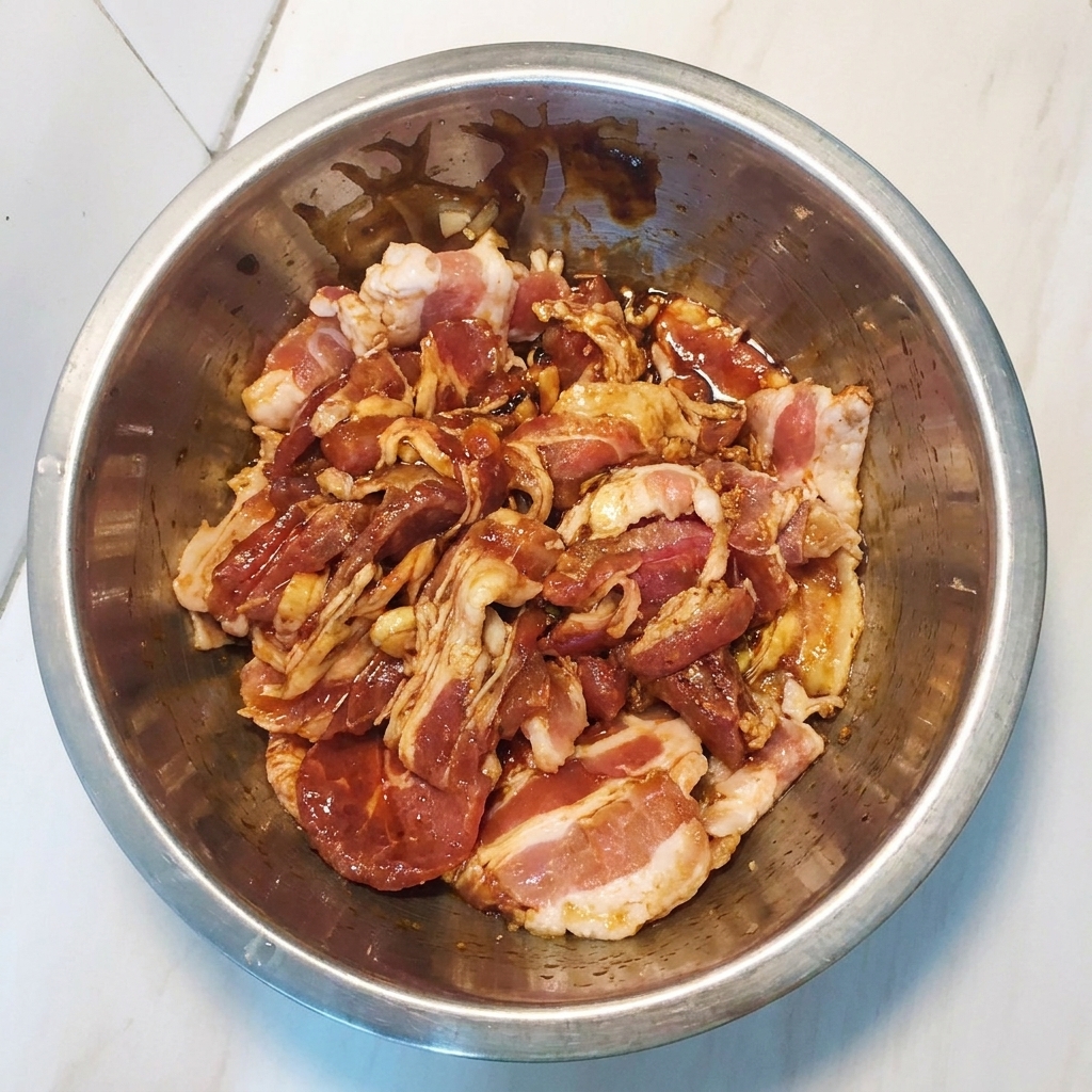 Rodajas de panceta de cerdo marinada siendo mezcladas en un bol de metal para asegurar una cobertura uniforme.