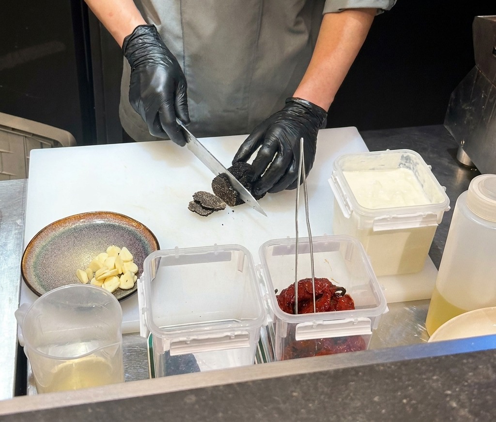 Un chef con guantes negros picando chiles negros secos en una tabla de cortar blanca.