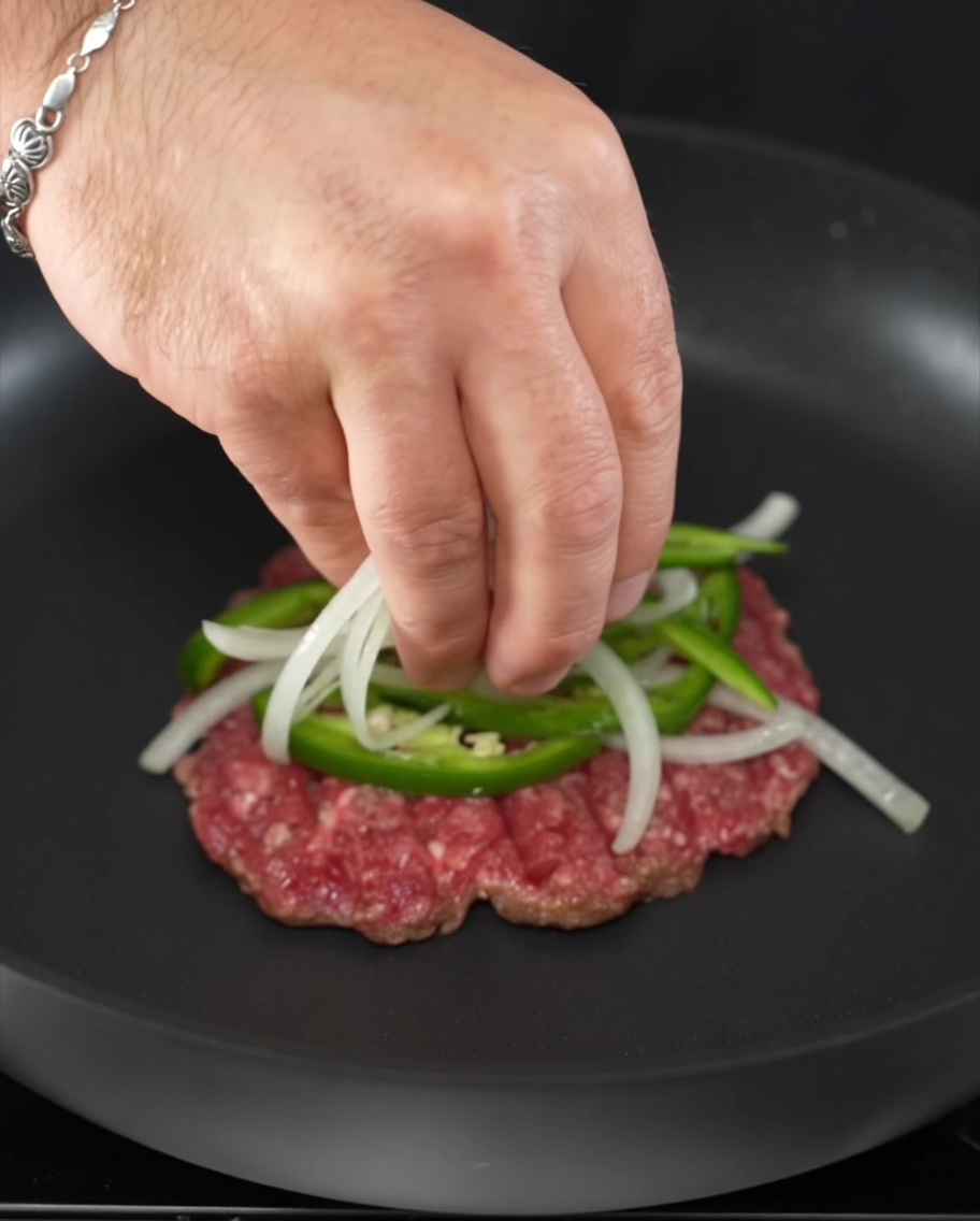 Rodajas de jalapeño fresco y cebolla colocadas encima de una hamburguesa chisporroteante.