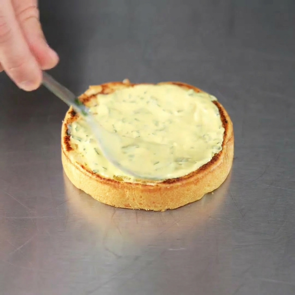 Un pan de hamburguesa tostado siendo untado con salsa verde, listo para la carne.