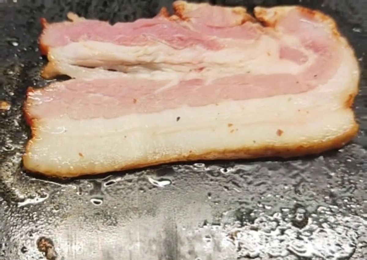Una tira de beicon chisporroteando y friéndose sobre la superficie caliente y plana de una plancha.