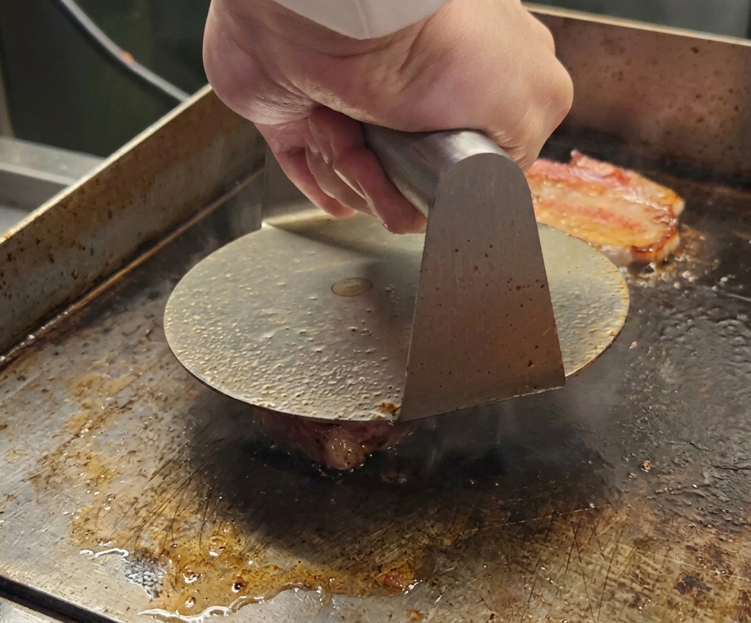 Una prensa de metal aplastando una bola de carne Wagyu picada sobre una superficie de cocción caliente.