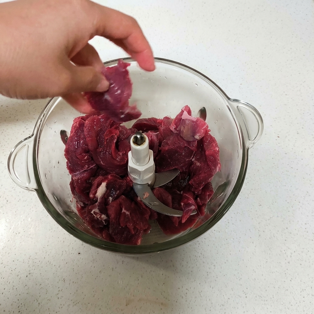 Una mano colocando trozos de carne cruda en el procesador de alimentos.