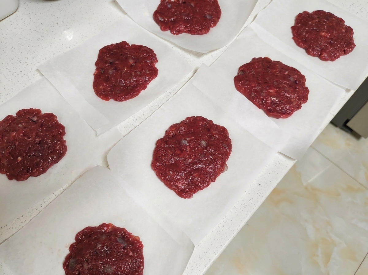 Varias hamburguesas crudas sobre cuadrados de papel de horno en una encimera.