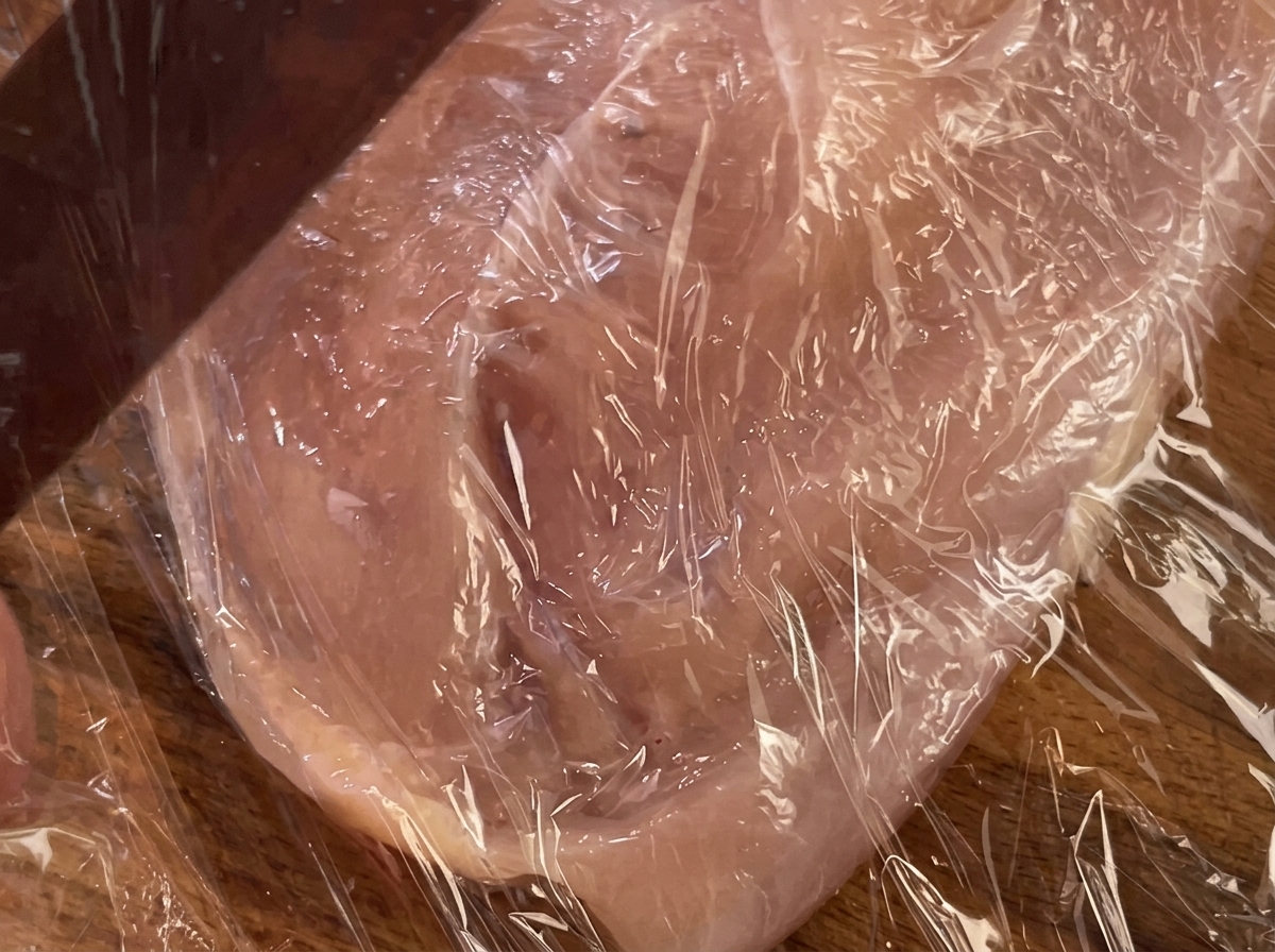 Un rodillo presionando una pechuga de pollo cubierta con film transparente.