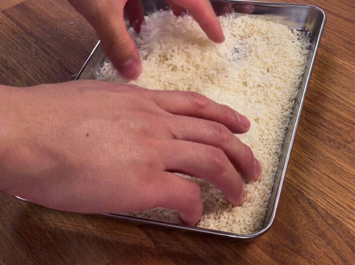 Manos presionando una pechuga de pollo en una bandeja de pan rallado y queso parmesano.