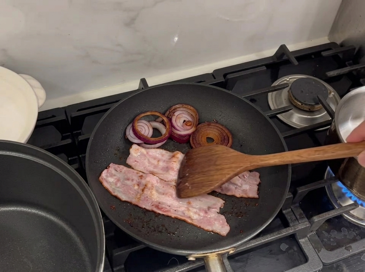 Tiras de tocino y aros de cebolla morada chisporroteando en una sartén.