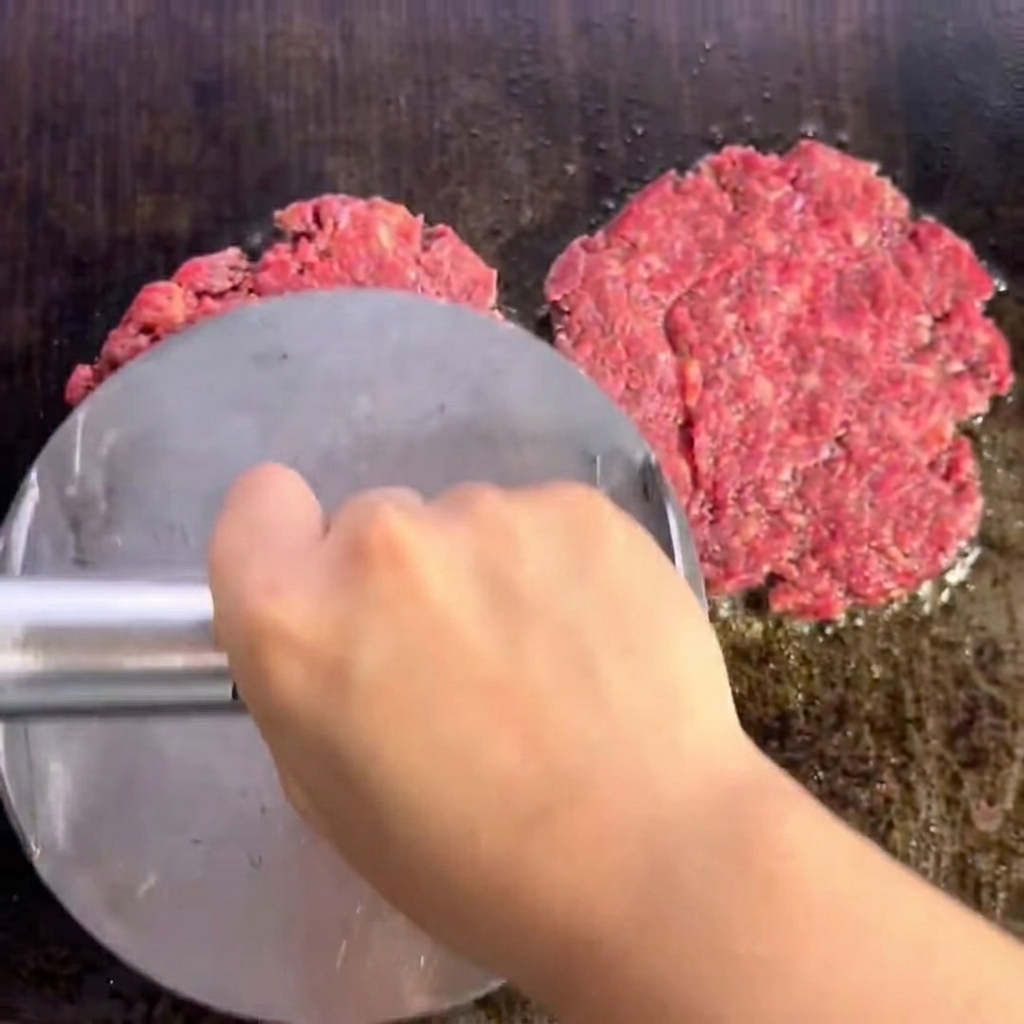 Dos hamburguesas de ternera sazonadas chisporroteando en la plancha, mostrando una costra dorada y sellada.