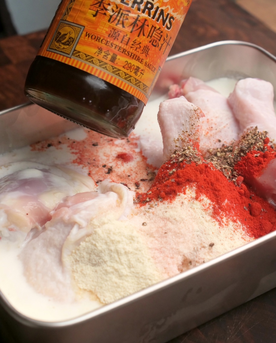 Salsa Worcestershire goteando sobre una bandeja de muslos de pollo crudos marinados y cubiertos con especias en polvo rojas.