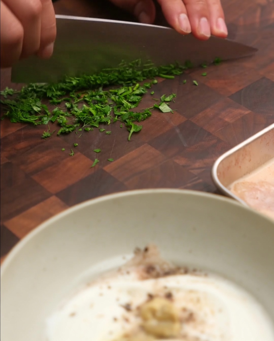 Cebollino y perejil frescos picados siendo añadidos a un bol de mezcla con yogur y otros ingredientes de la salsa.