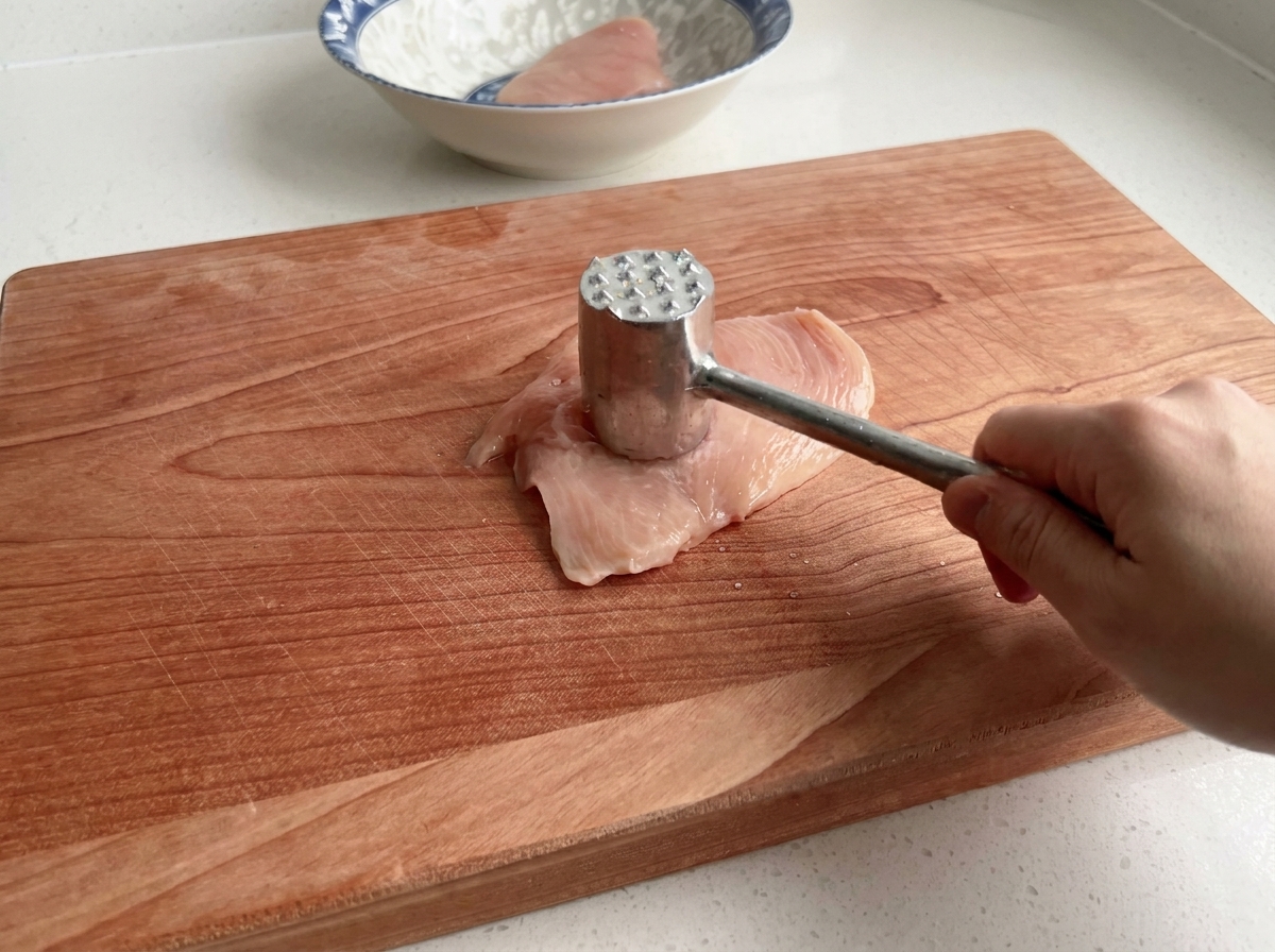 Primer plano de un mazo de cocina ablandando una pechuga de pollo cruda sobre una superficie de madera.