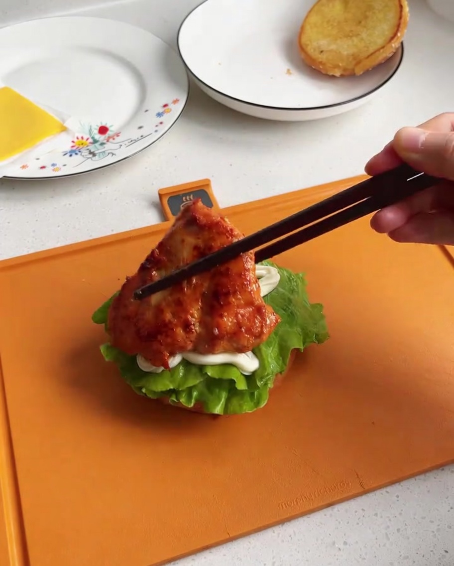 Un trozo de pollo cocinado y glaseado siendo colocado sobre la base de la hamburguesa con lechuga y mayonesa.