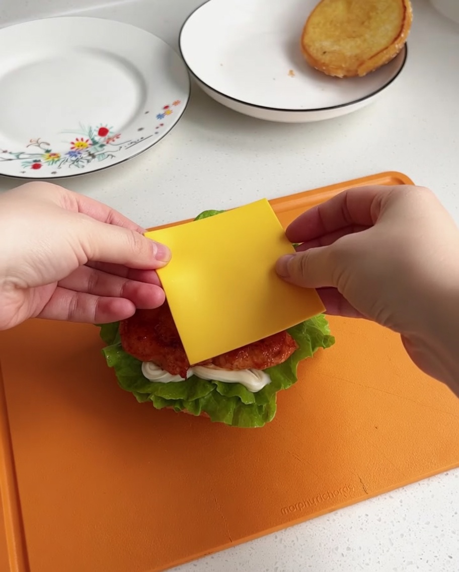 Una loncha de queso amarillo siendo centrada sobre el trozo de pollo caliente en la base de la hamburguesa.
