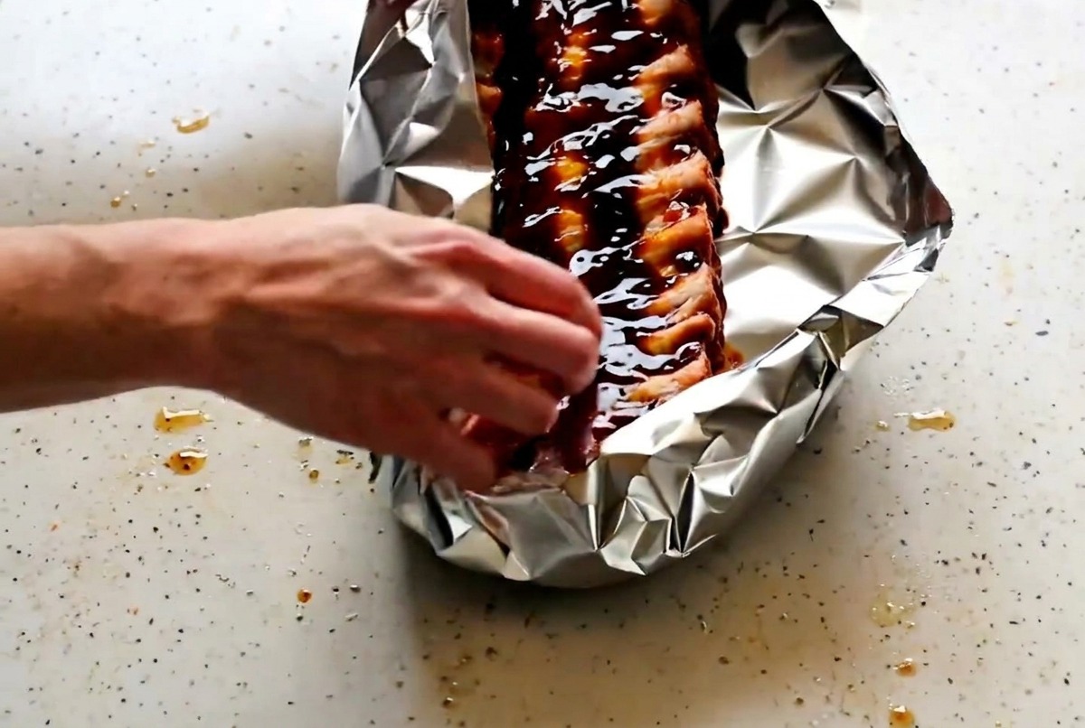Manos doblando y sellando cuidadosamente los bordes del papel de aluminio para cerrar las costillas.
