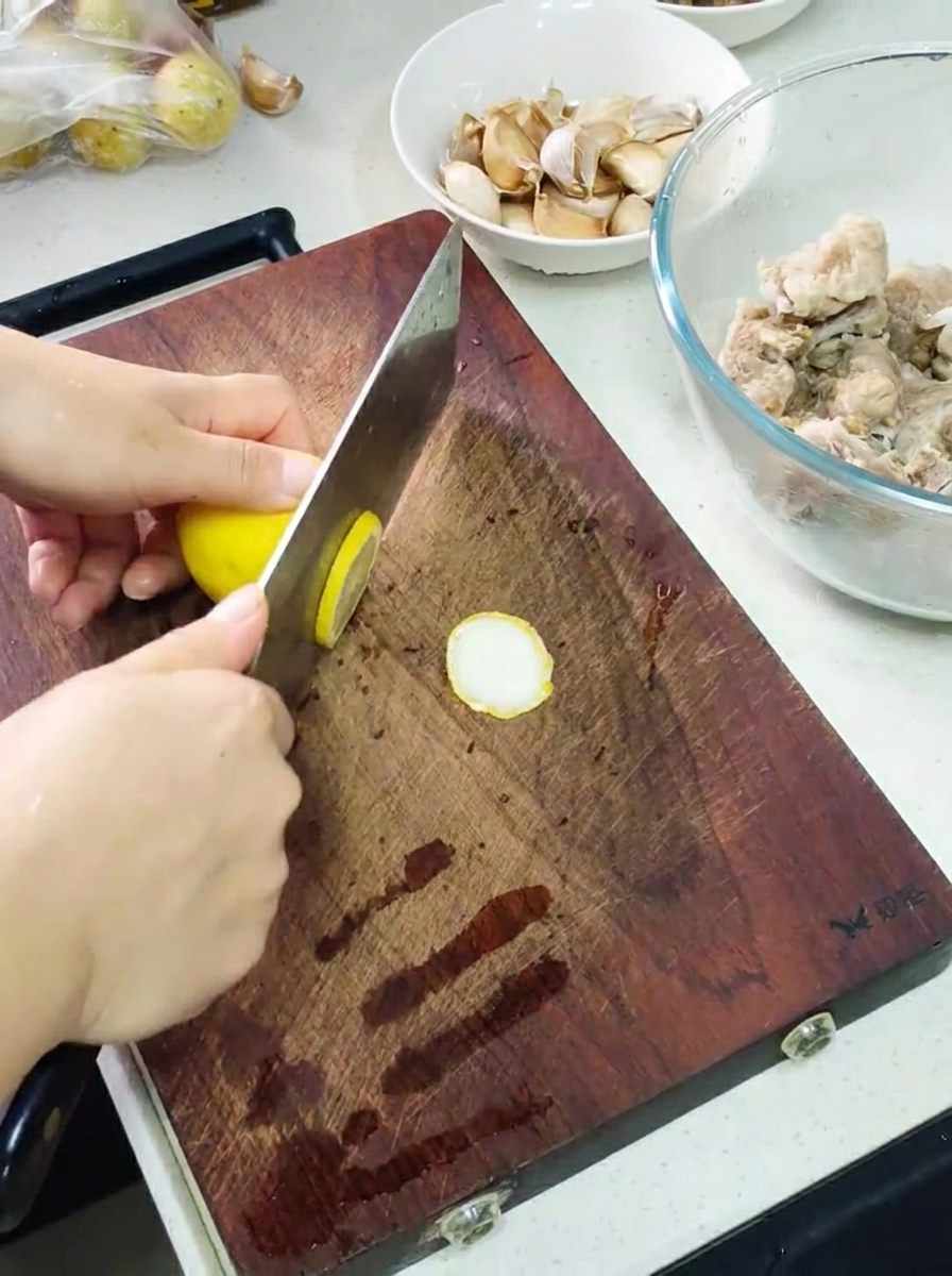 Manos usando un cuchillo para rebanar un limón sobre una tabla de cortar de madera, con un bol de costillas marinadas y dientes de ajo cerca.