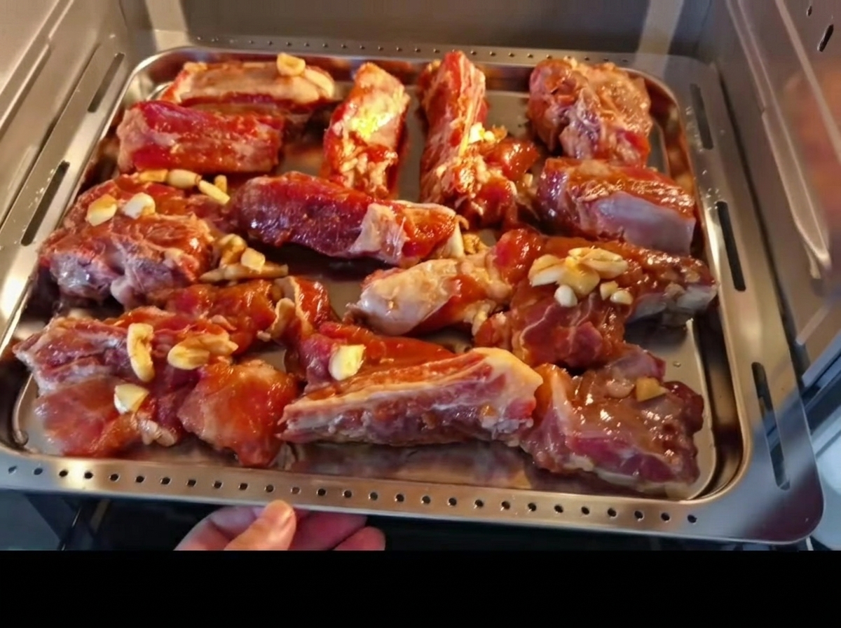 Costillas de cerdo marinadas dispuestas uniformemente en una bandeja de metal, siendo colocadas en un horno.