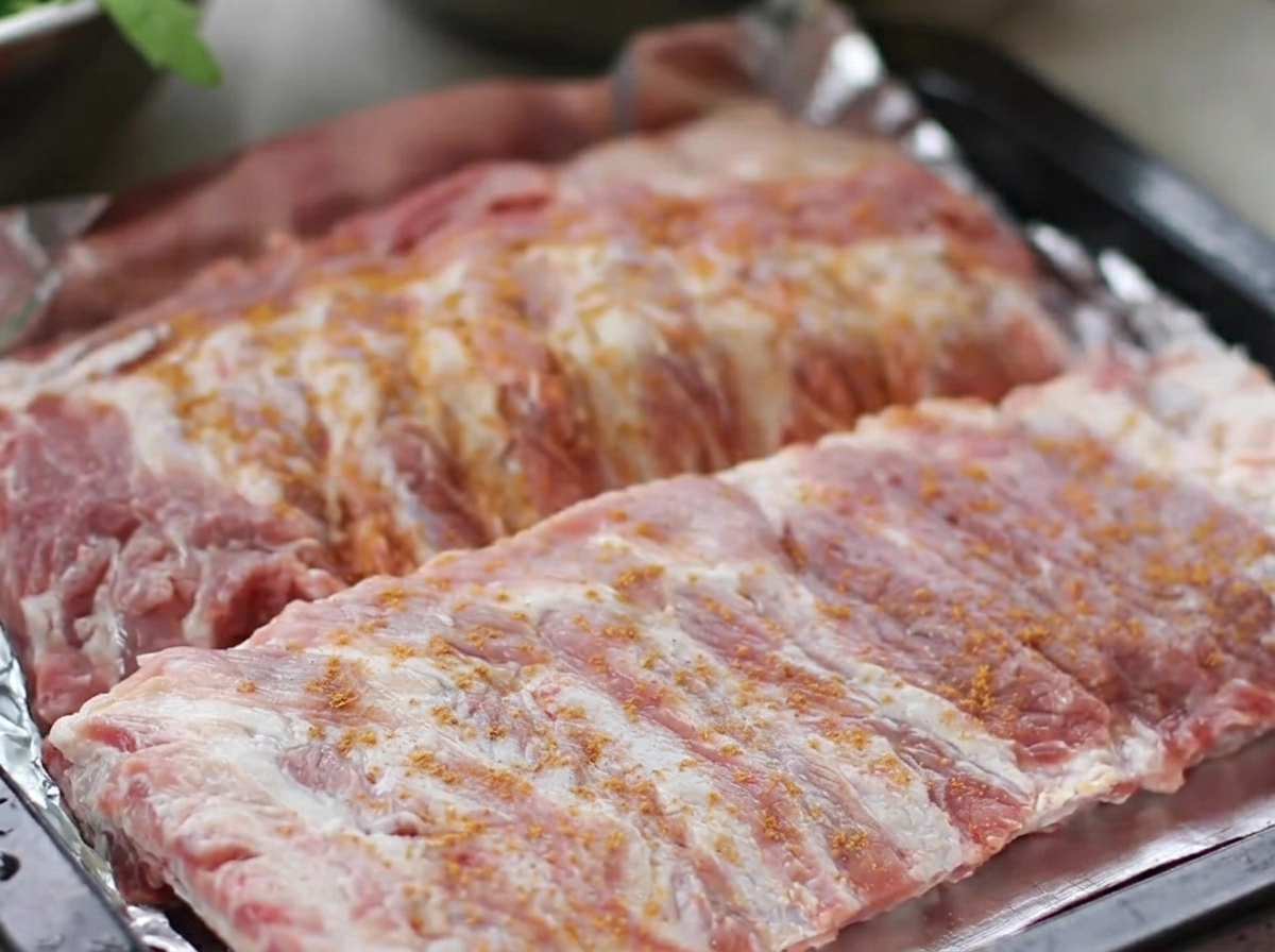Costillas de cerdo crudas dispuestas en una bandeja de horno, generosamente cubiertas con un aliño seco de cinco especias, pimienta negra, jengibre y ajo.