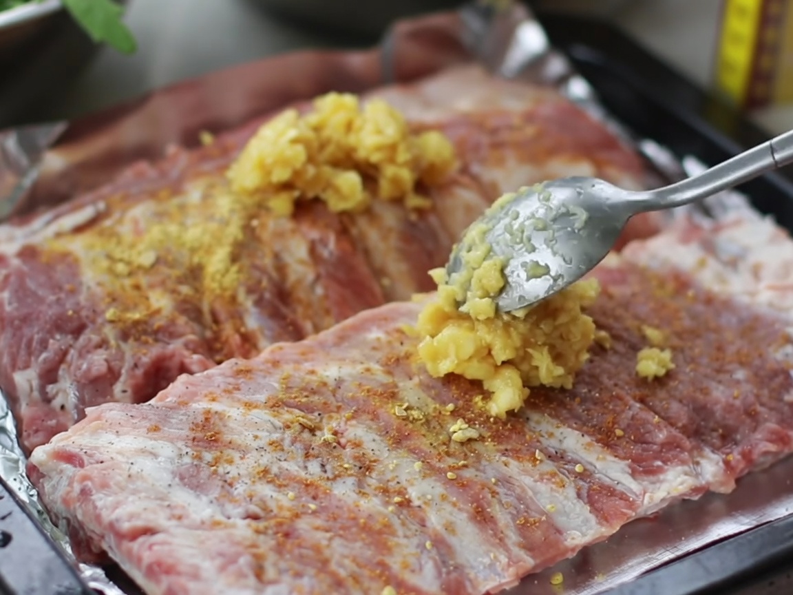 Manos frotando una mezcla de ajo picado y especias sobre la superficie de costillas de cerdo crudas en una bandeja.