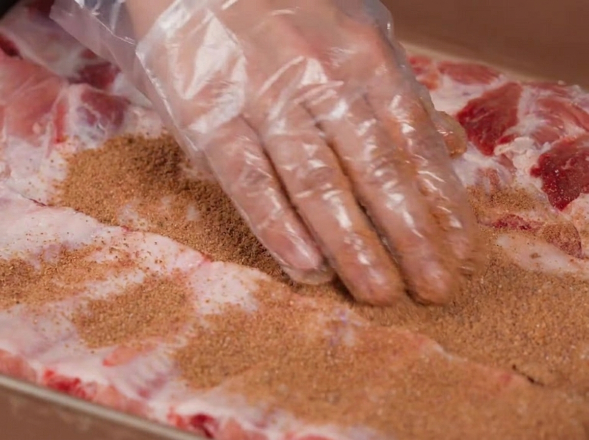Manos usando guantes de plástico frotando una mezcla de especias marrón sobre costillas de cerdo crudas.