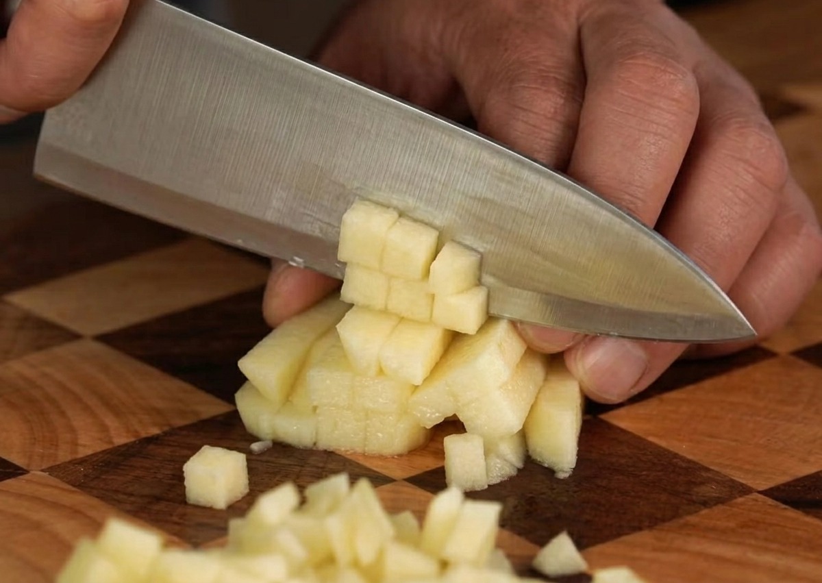 Una manzana fresca siendo cortada cuidadosamente en pequeños cubos uniformes sobre una tabla de madera.